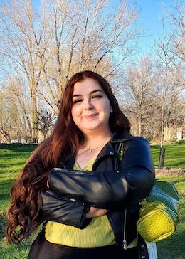 Marili, wearing a green shirt and leather jacket, standing in a park with her arms crossed