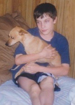 A young Patrick in a blue shirt and black short sitting on his bed holding a dog